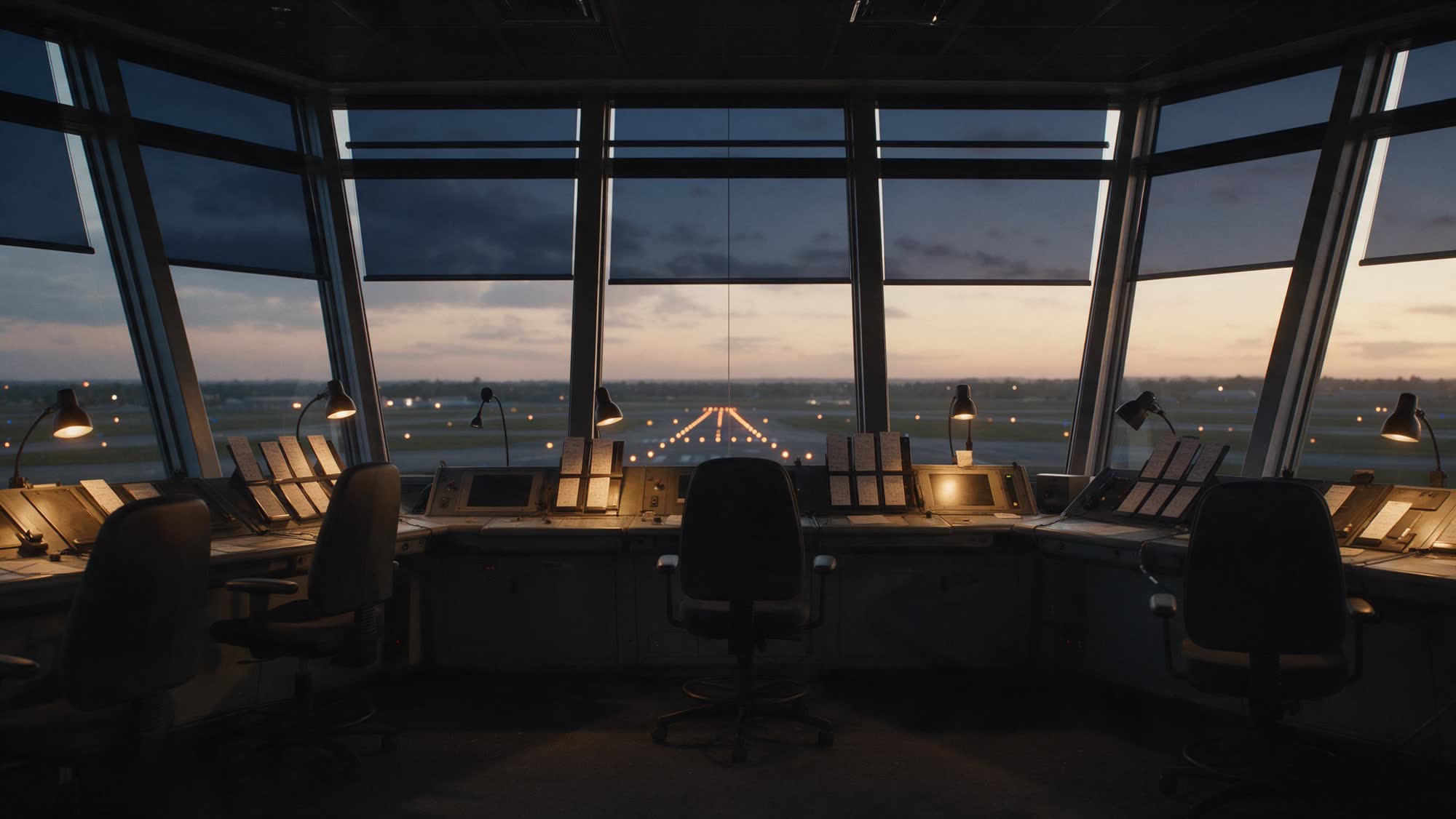 An empty control tower cab at dusk, paper flight strips in their holders, runway lights beyond the glass